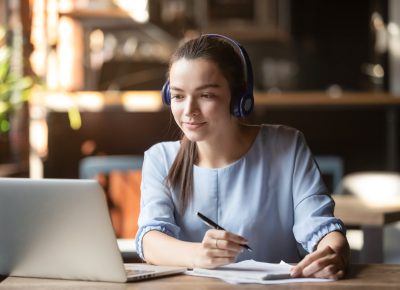 Focused,Woman,Wearing,Headphones,Using,Laptop,In,Cafe,,Writing,Notes,